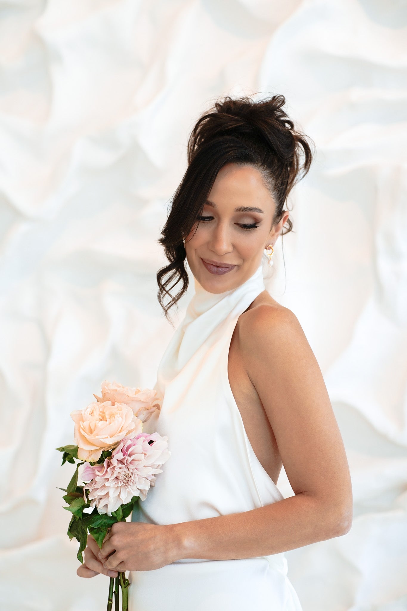 Bride in white halter dress smiling with eyes closed and holding bouquet – wearing gold Sophia Earrings against soft white backdrop