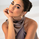 Bridesmaid wearing Jayda Earrings with dark hair in bun and purple halter dress – leaning head on palm, curious expression against white background