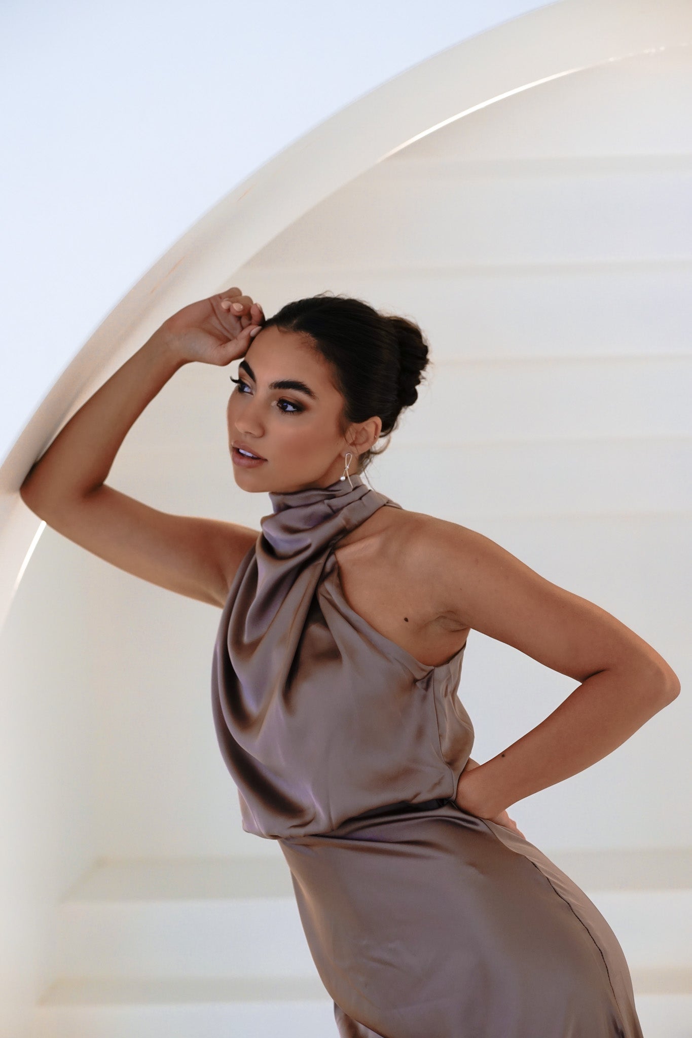 Woman wearing Kiera Earrings as a bridesmaid in a purple-grey halter dress, hair in a bun, leaning on an arched wall with head resting on hand