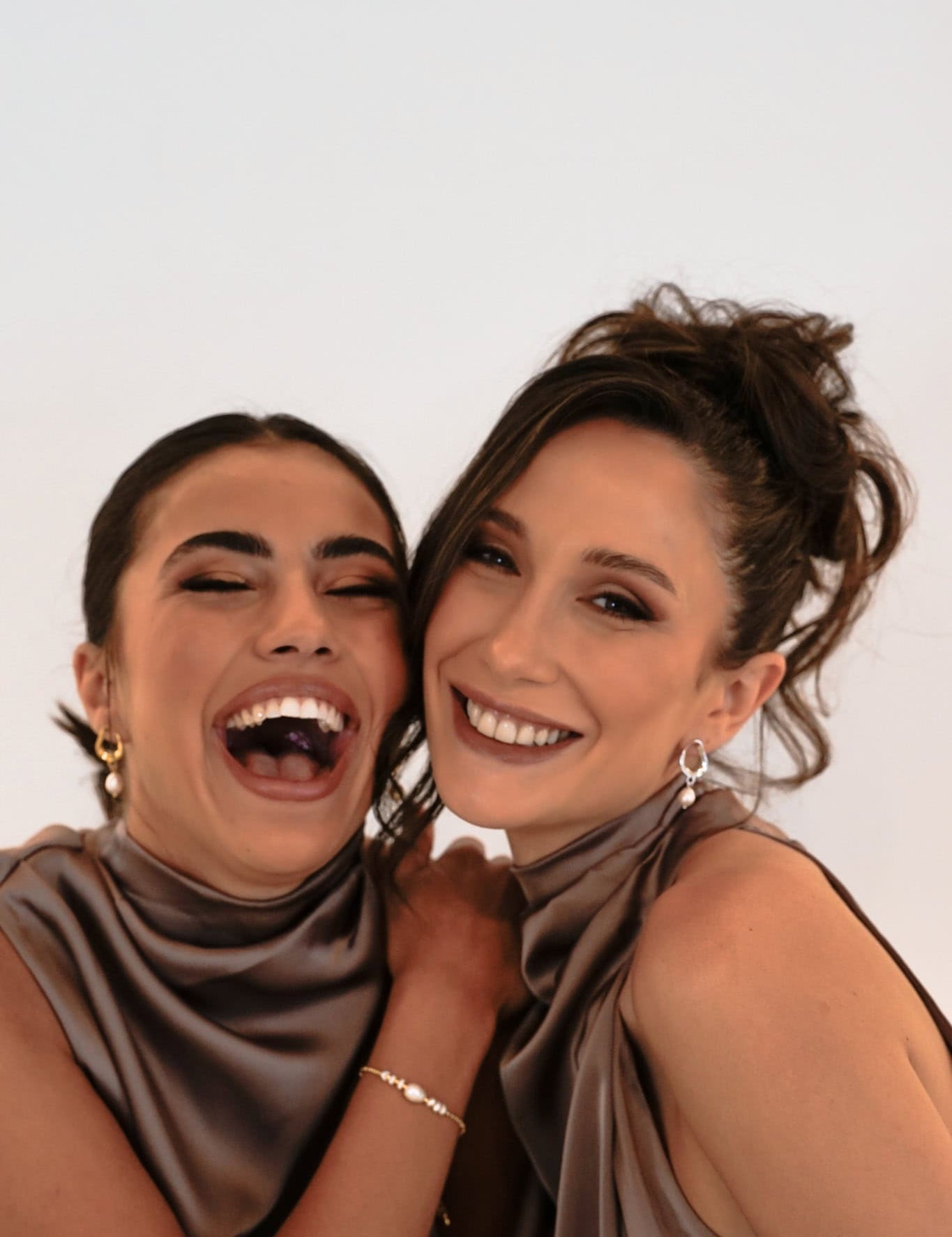 Two bridesmaids in purple dresses, smiling and laughing – one wears gold Sophia Earrings, the other silver, both with updos against a white backdrop