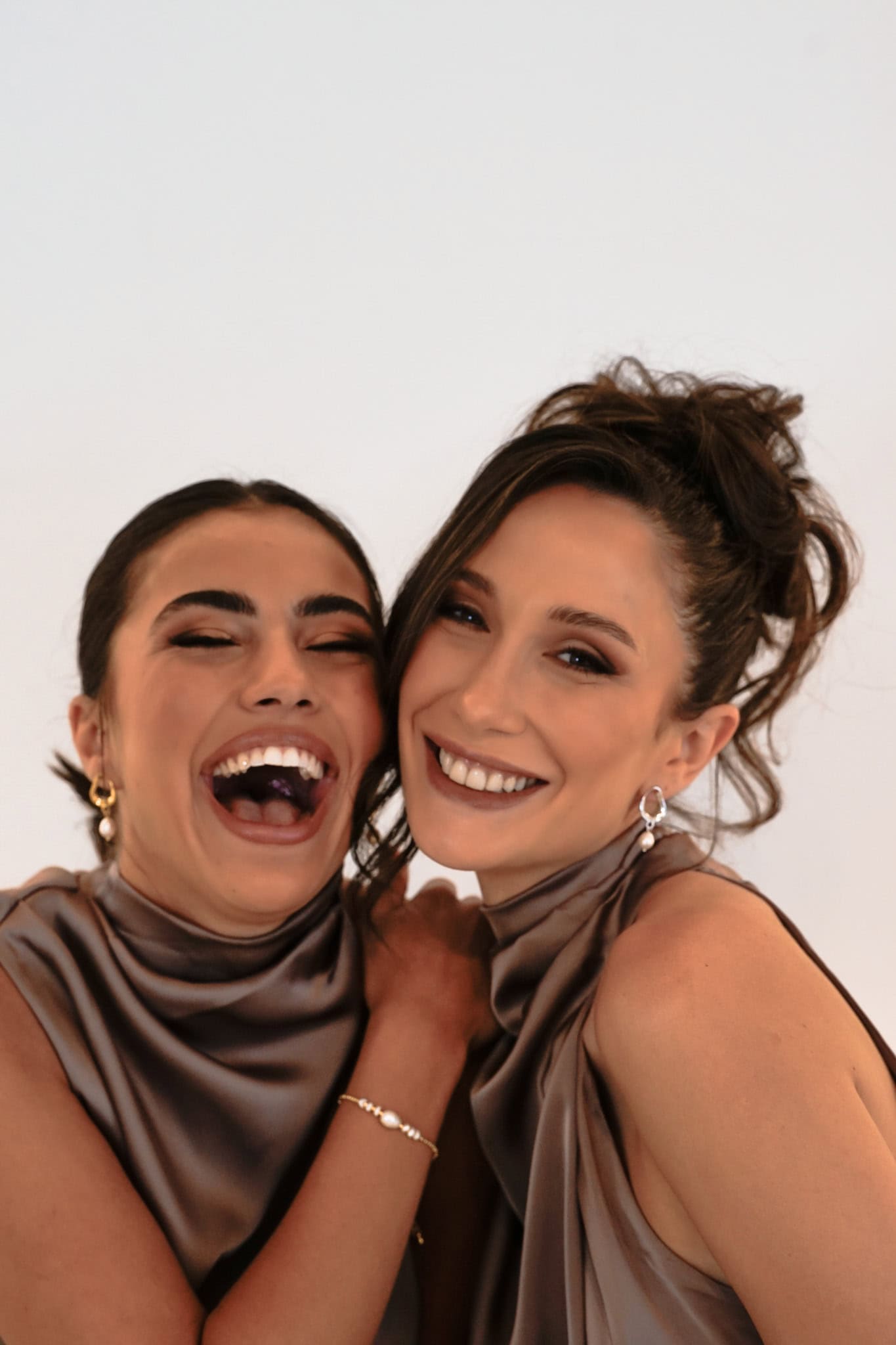 Two bridesmaids in purple dresses, smiling and laughing – one wears gold Sophia Earrings, the other silver, both with updos against a white backdrop