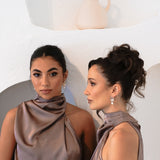 Two bridesmaids in purple halter dresses wearing Jayda Earrings – one looking into camera, the other in profile near a soft ceramic pitcher on a stucco alcove shelf