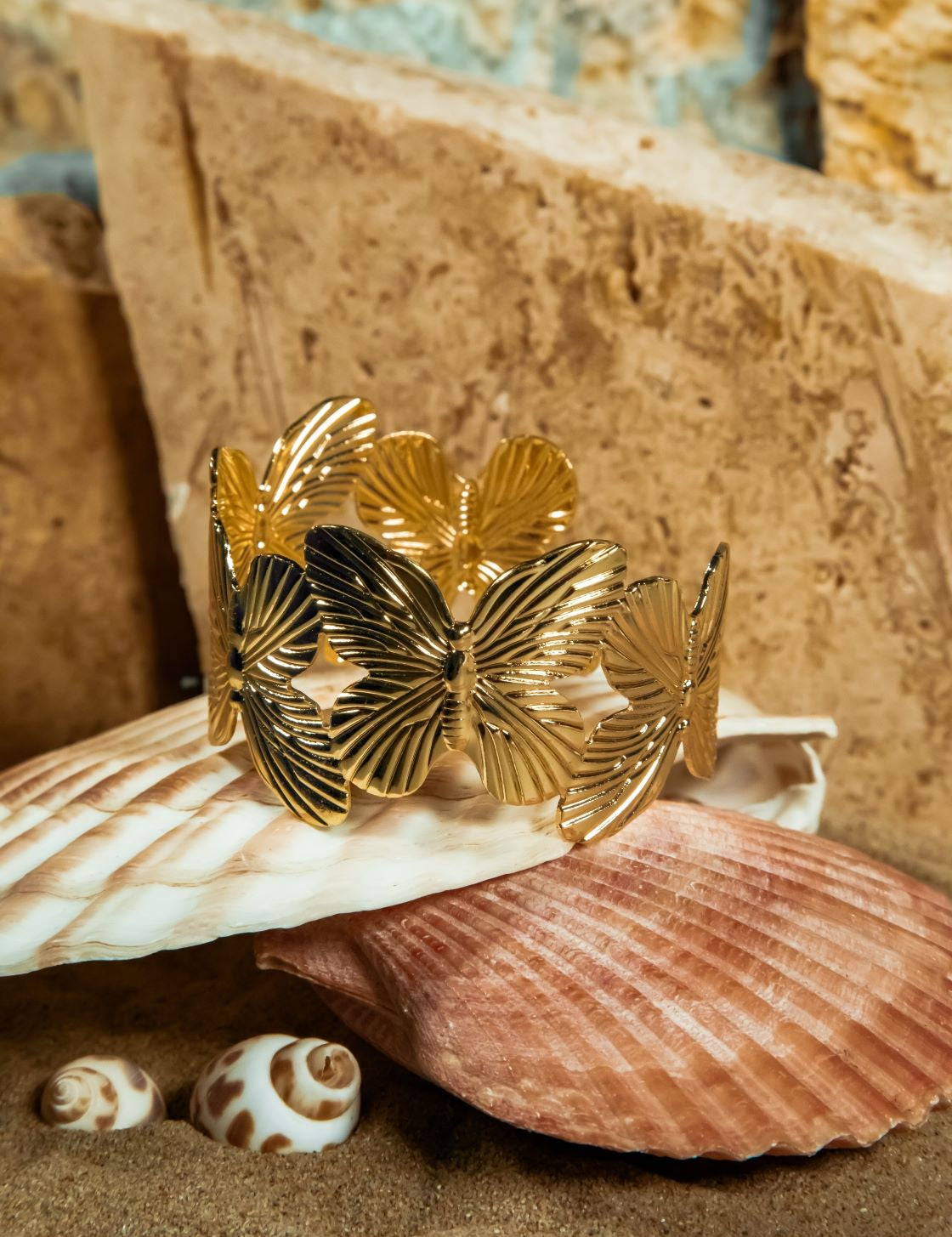 Gold Gia Bracelet displayed atop a closed clamshell on beach sand – textured sandstone brick softly blurred in the background