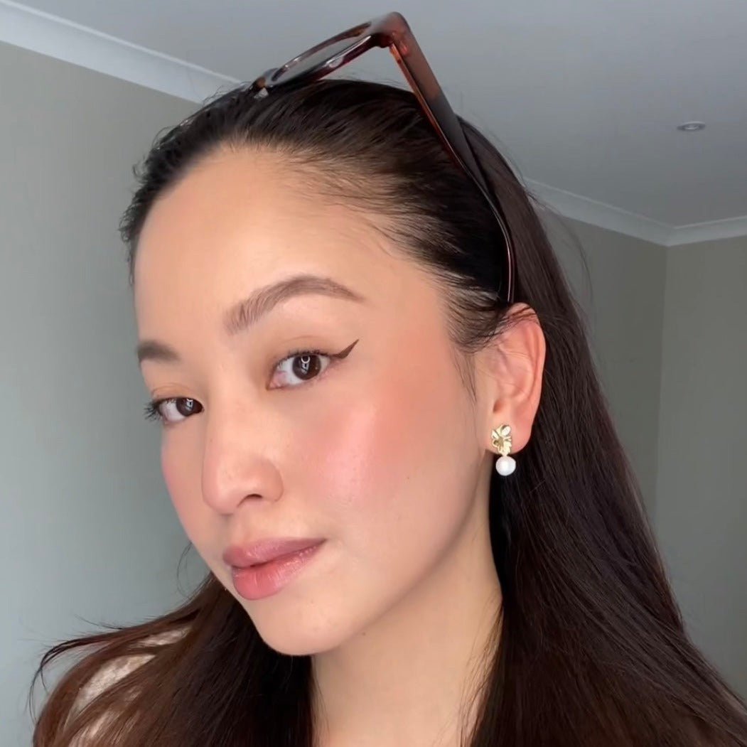 Woman wearing Mini Destiny Earrings in gold – straight brunette hair, sunglasses atop head, close-up selfie highlighting pearl detail