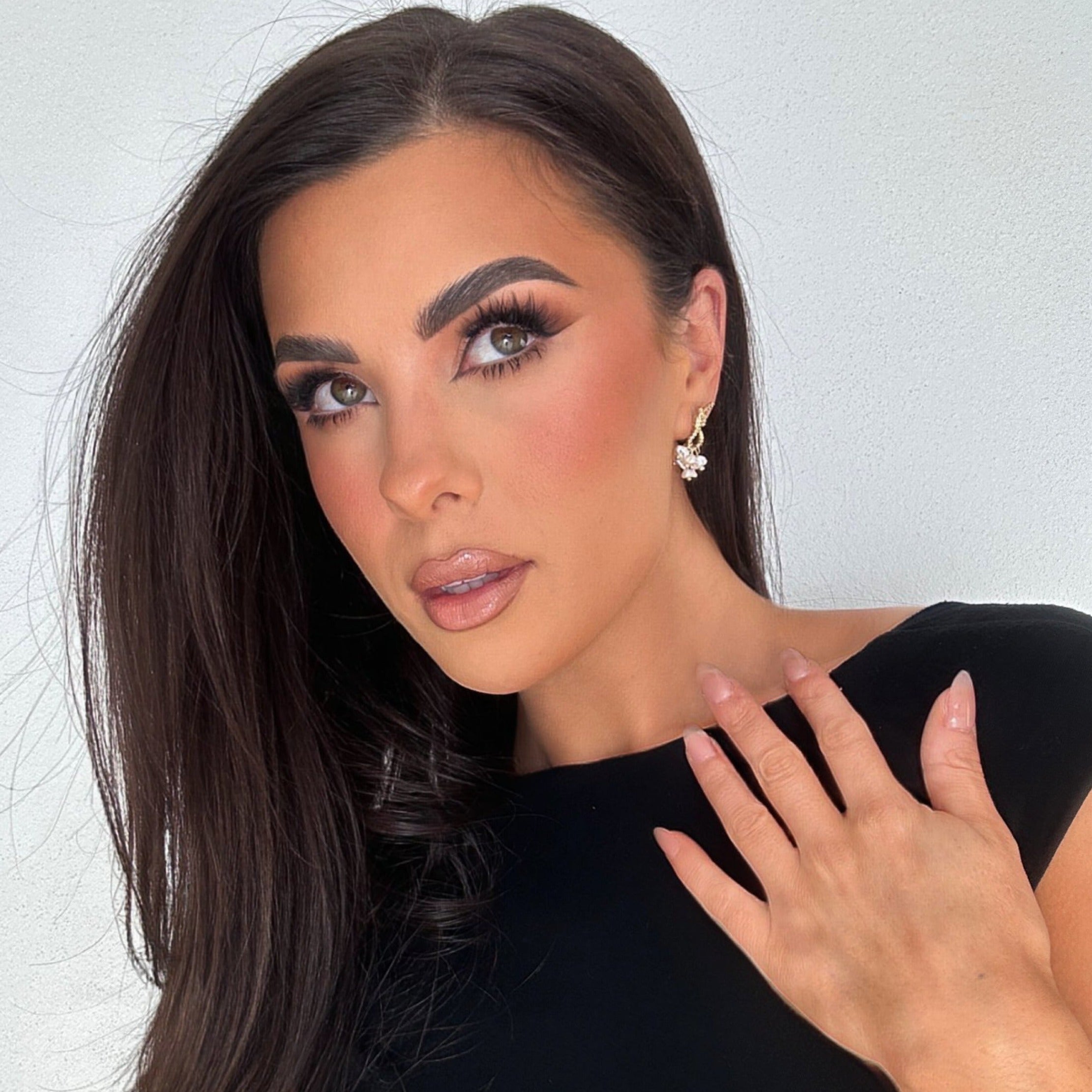 Woman with dark flowing hair and glam makeup wearing Loretta Earrings – posed with hand near shoulder, gazing upward against soft off-white background