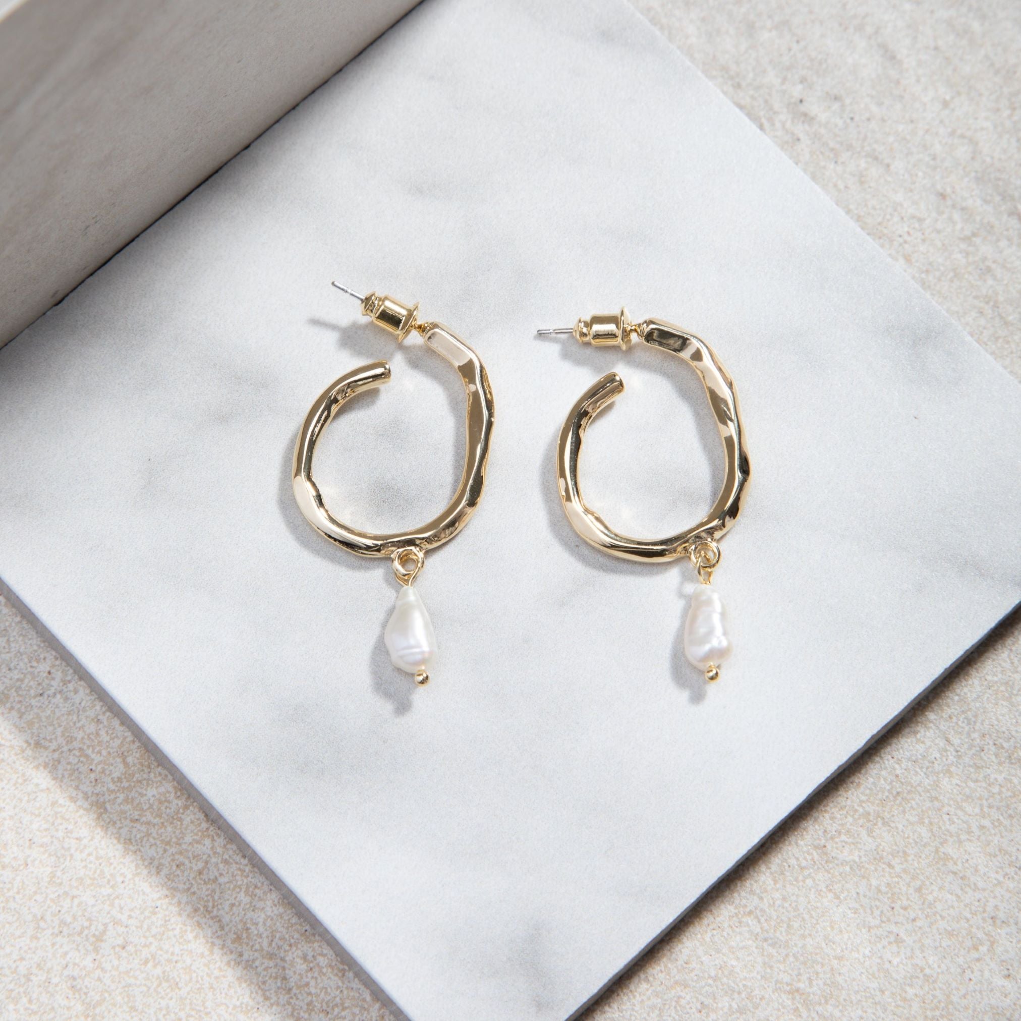 Jolene Earrings in gold – freshwater pearl hoops styled on white tile with mottled sand-toned felt backdrop