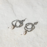 Isabella Earrings – silver twisted hoop earrings with suspended pearl, resting on matte white tile surface