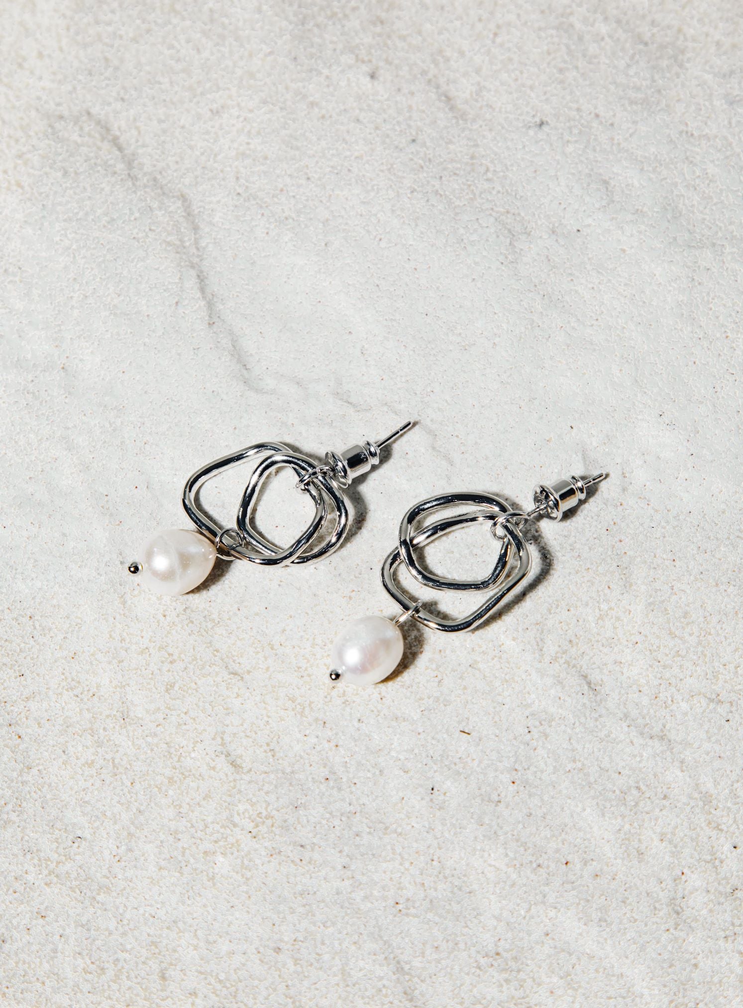 Isabella Earrings – silver twisted hoop earrings with suspended pearl, resting on matte white tile surface