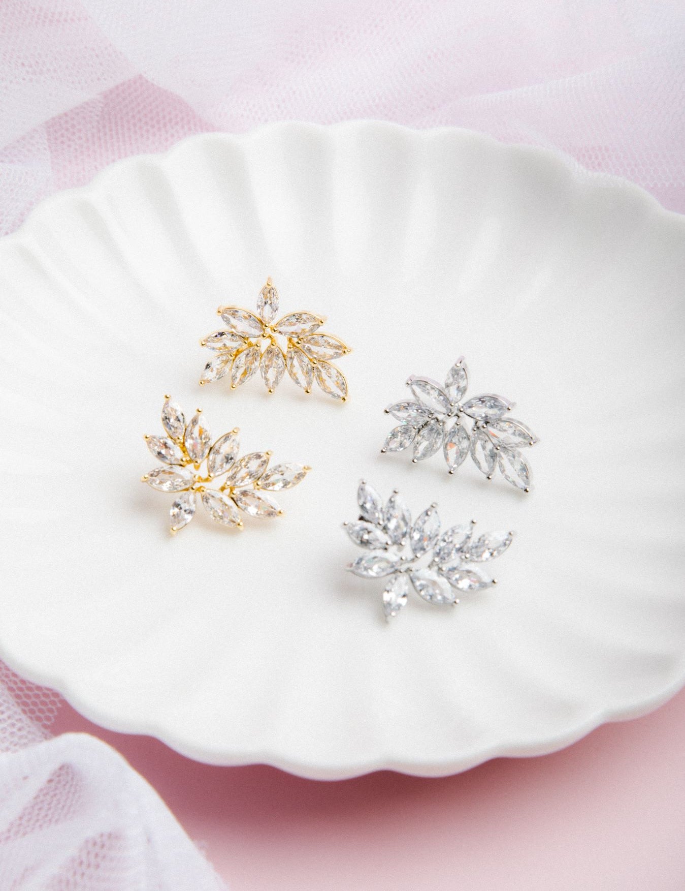 Kya Earrings in gold and silver displayed side by side on a scalloped porcelain plate with a soft pink glow and delicate veil in the background
