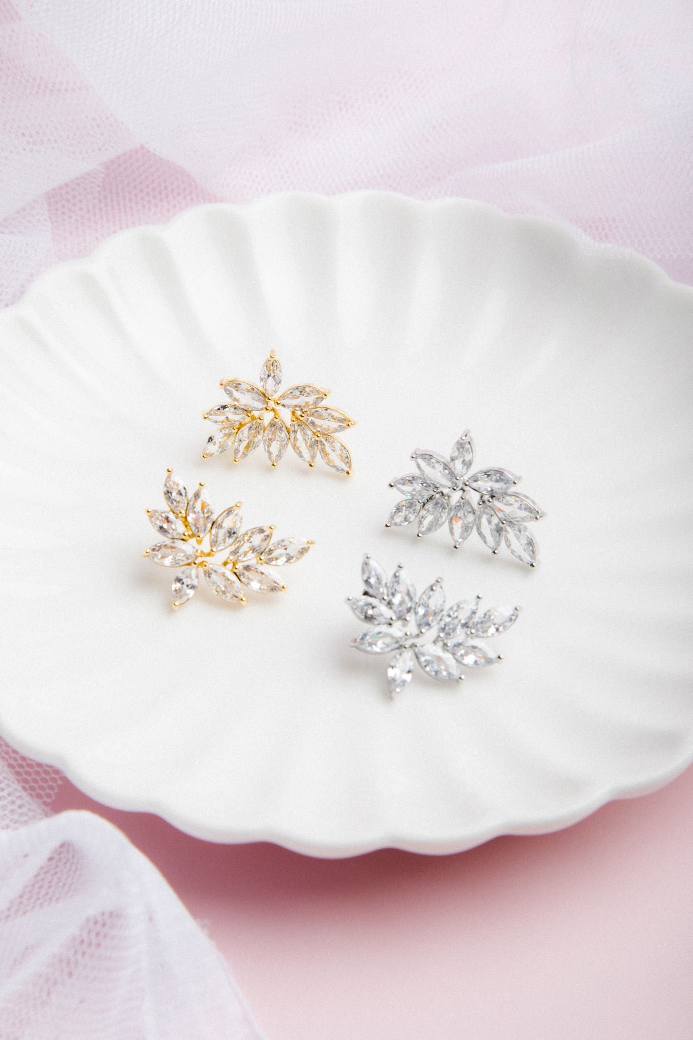 Kya Earrings in gold and silver displayed side by side on a scalloped porcelain plate with a soft pink glow and delicate veil in the background