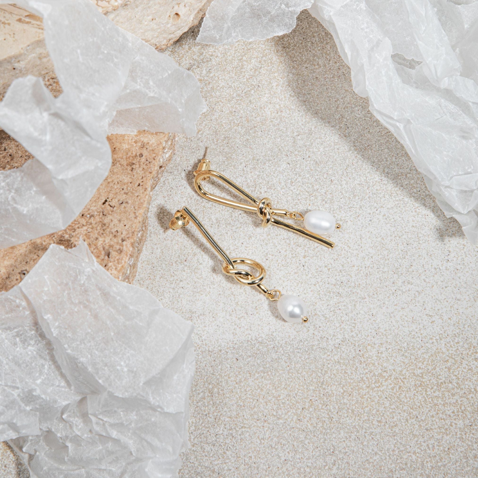 Syntyche Earrings in gold flatlay on textured sandstone-grey surface, placed beside a brown sandstone paver with scattered wrapping paper