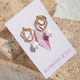 Isabella Earrings – gold twisted hoop earrings with pearl, displayed on Kymond & Co card with sandstone pavers in background