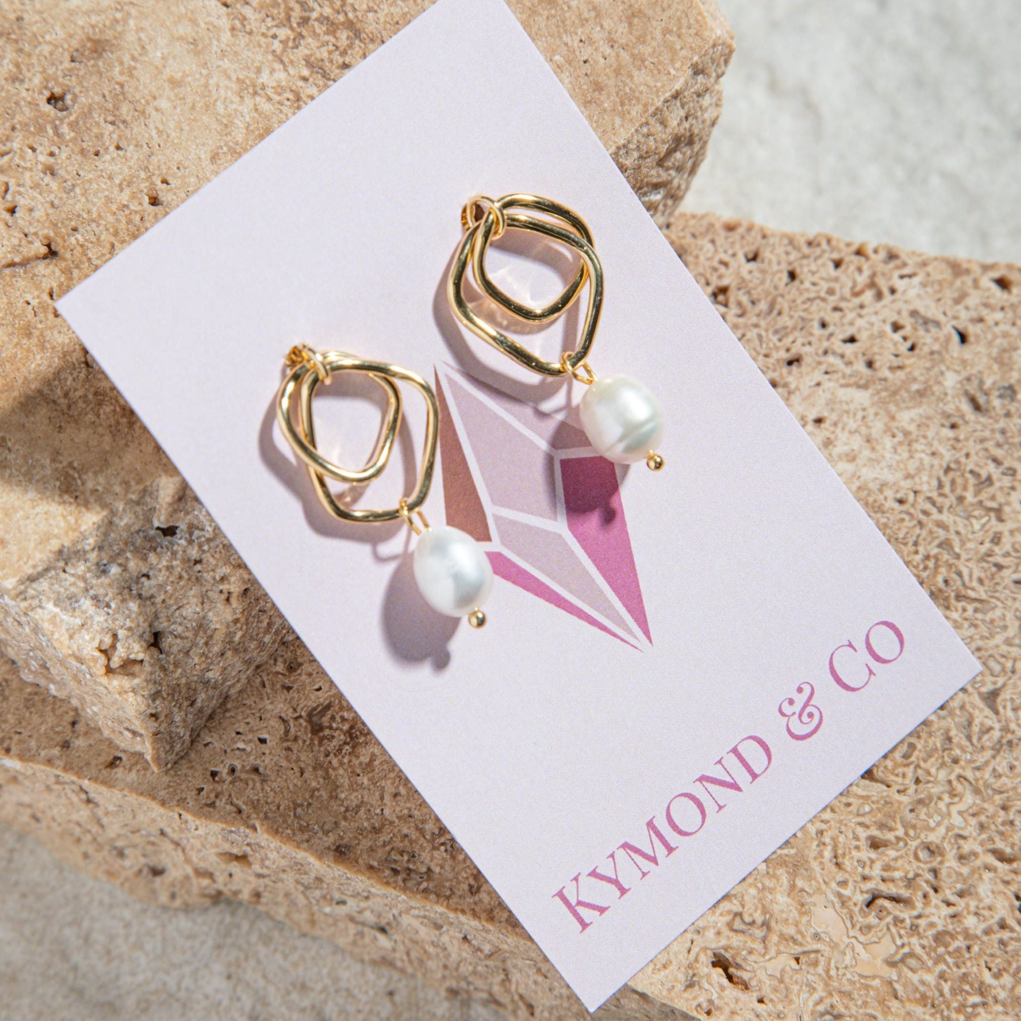 Isabella Earrings – gold twisted hoop earrings with pearl, displayed on Kymond & Co card with sandstone pavers in background