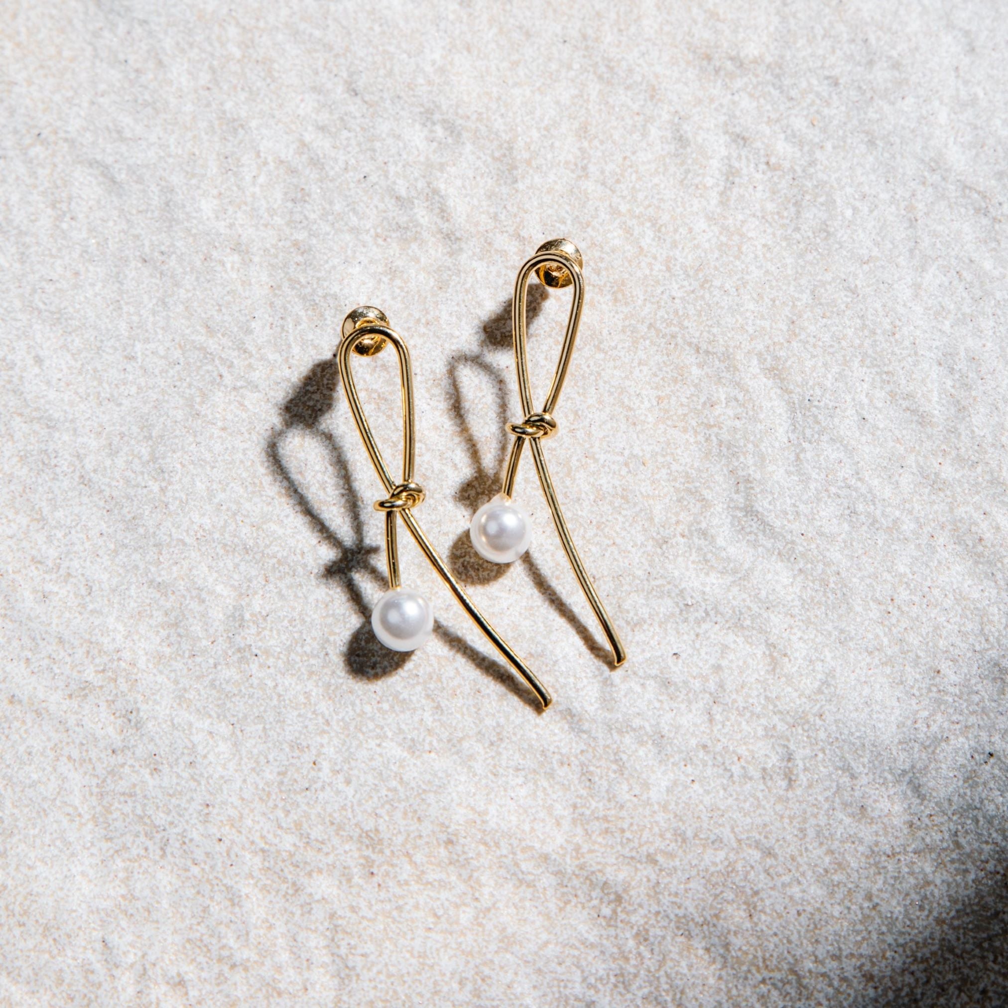 Kiera Earrings – gold dainty knot and pearl stud earrings on textured background