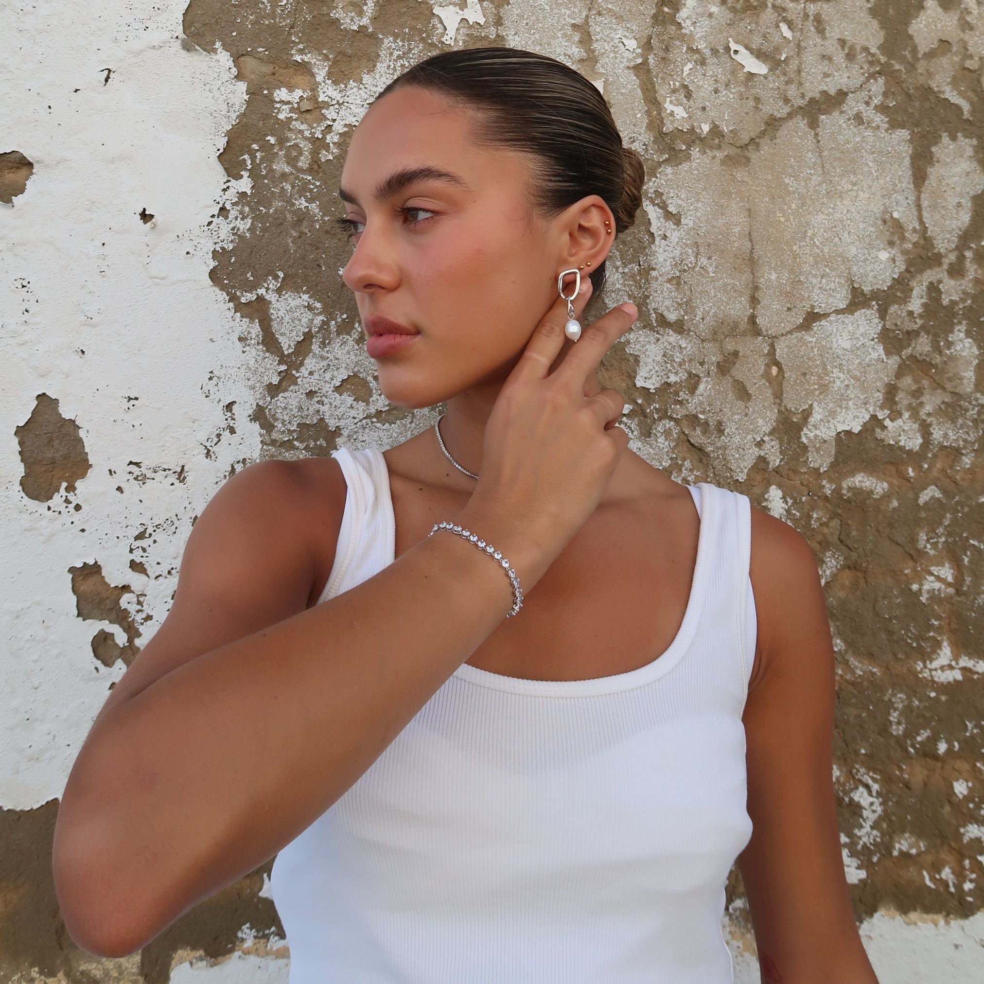 Woman wearing silver Sydney Earrings with hair in a bun, posing against textured white wall in a white tank top, hand behind ear to showcase earring