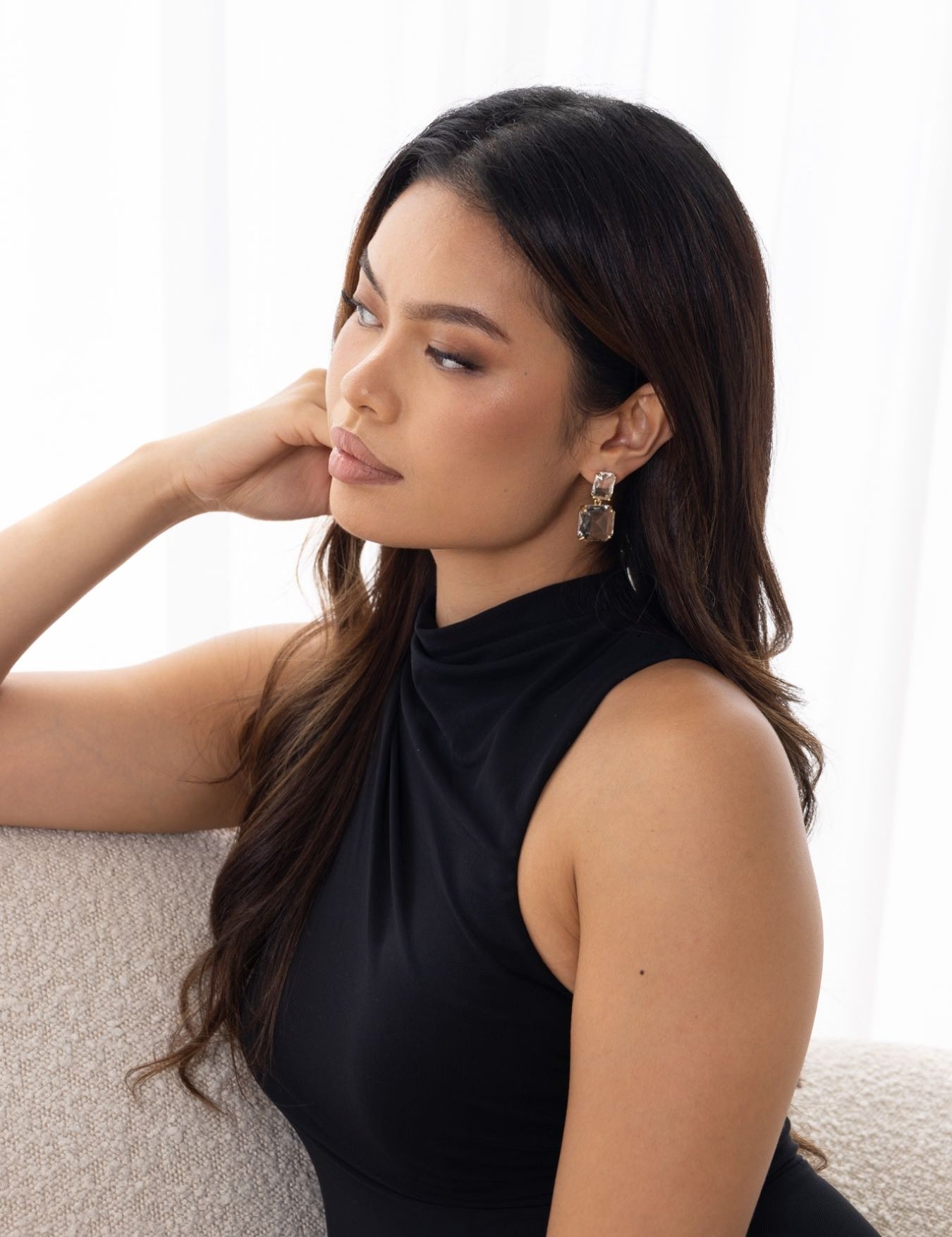 Woman in black dress seated on off-white couch, wearing Bianca Earrings – looking thoughtful with sheer curtains in bright background