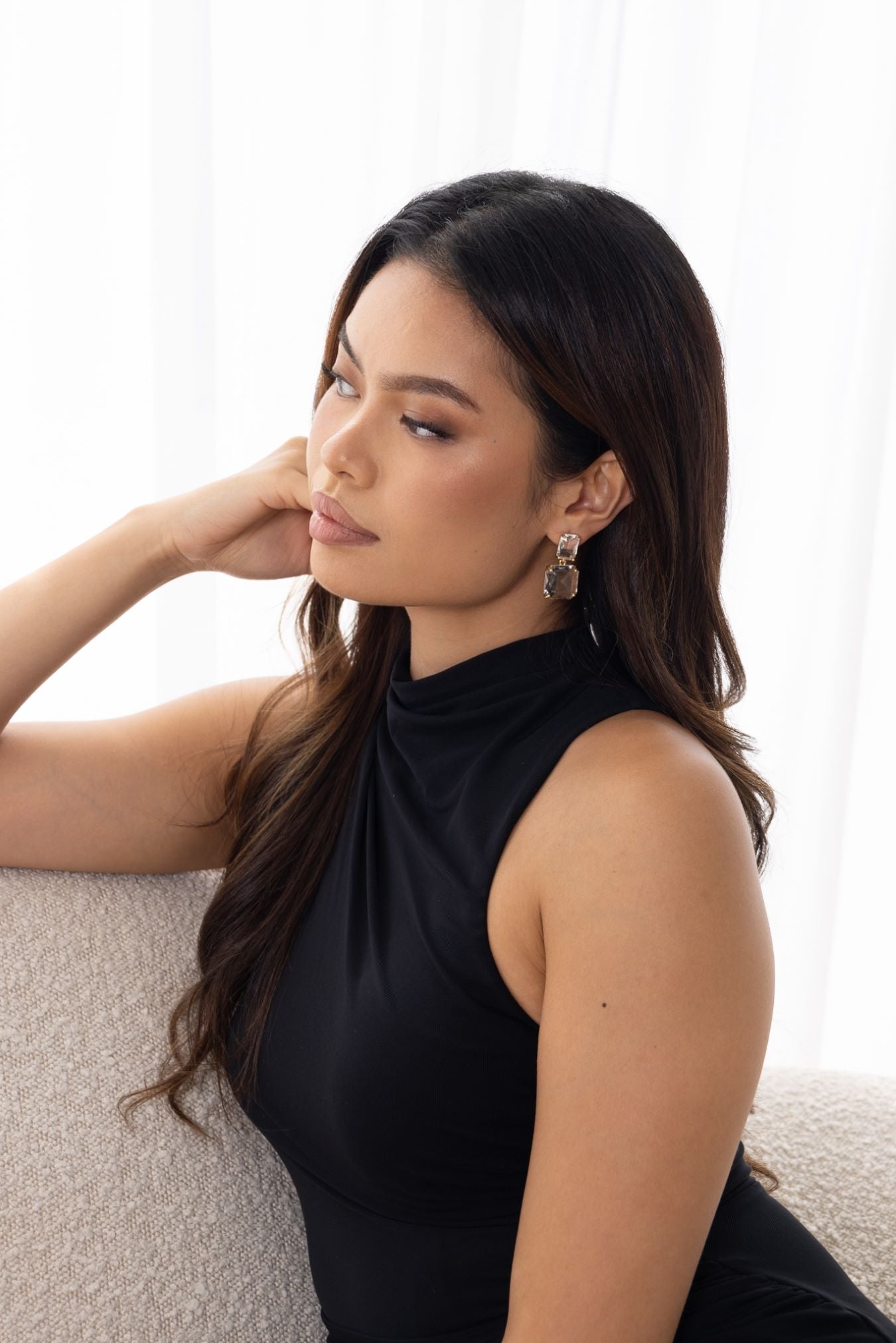 Woman in black dress seated on off-white couch, wearing Bianca Earrings – looking thoughtful with sheer curtains in bright background