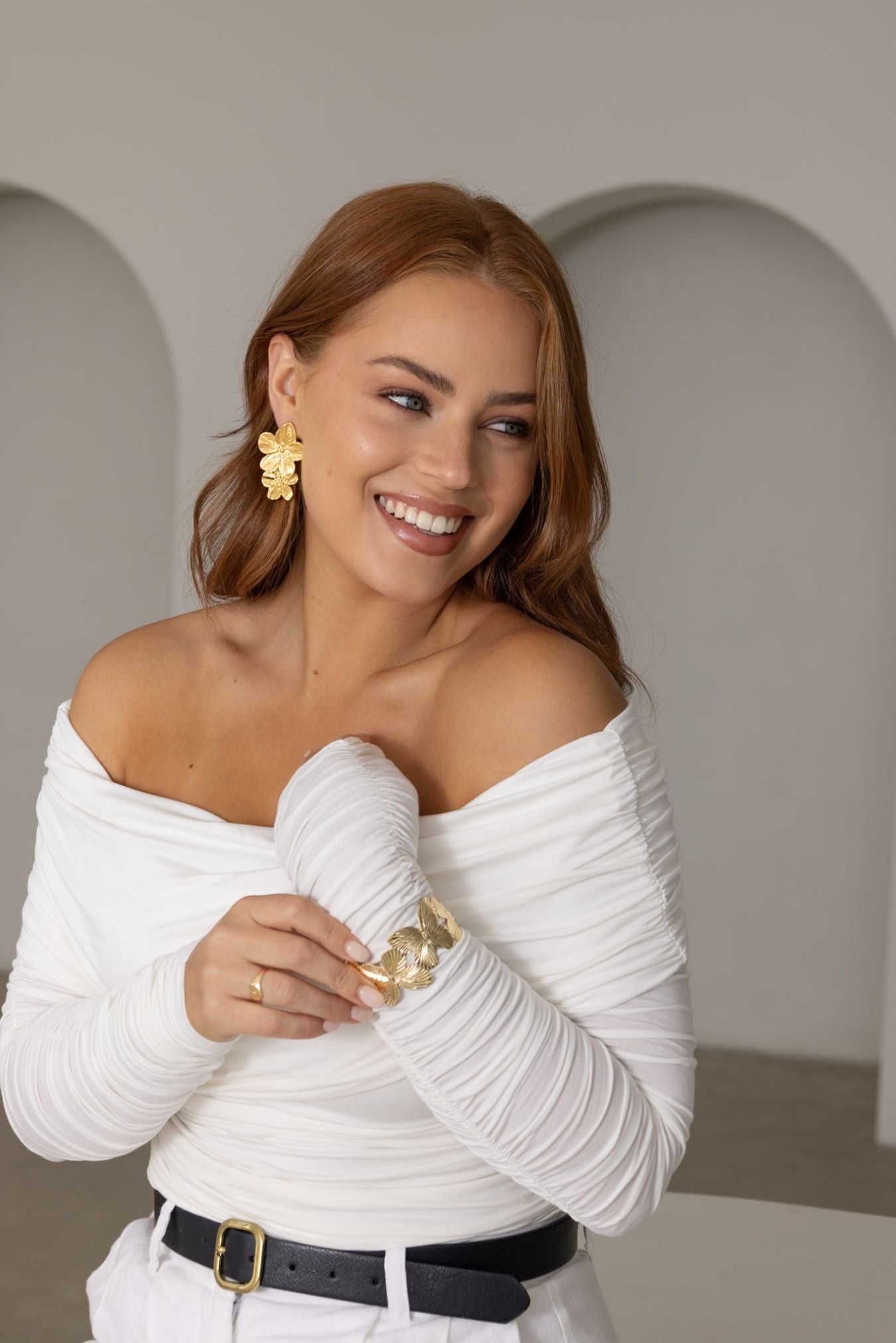Woman with copper brunette hair wearing Fleur Earrings – white off-shoulder blouse, gold cuff bracelet and matching accessories, smiling in beach house setting