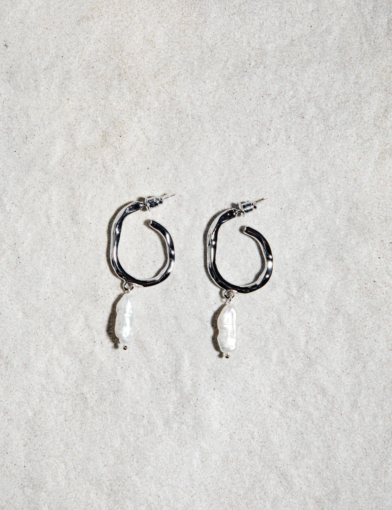 Jolene Earrings in silver – pearl drop hoops resting on softly textured sand-hued felt surface