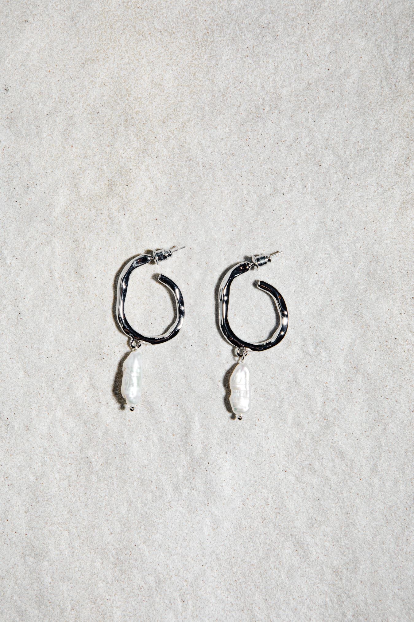Jolene Earrings in silver – pearl drop hoops resting on softly textured sand-hued felt surface