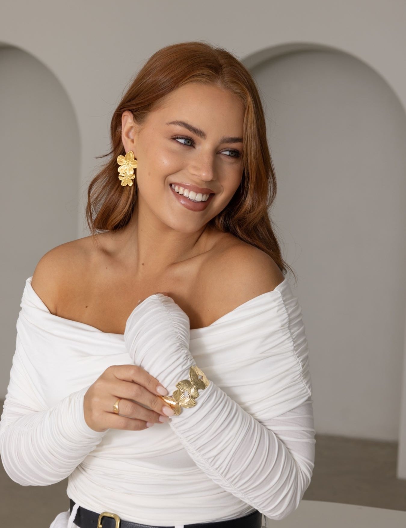 Woman with flowing copper hair wearing Gia Bracelet in gold – styled with white open-shoulder blouse, black belt and matching gold accessories, seated in soft coastal interior