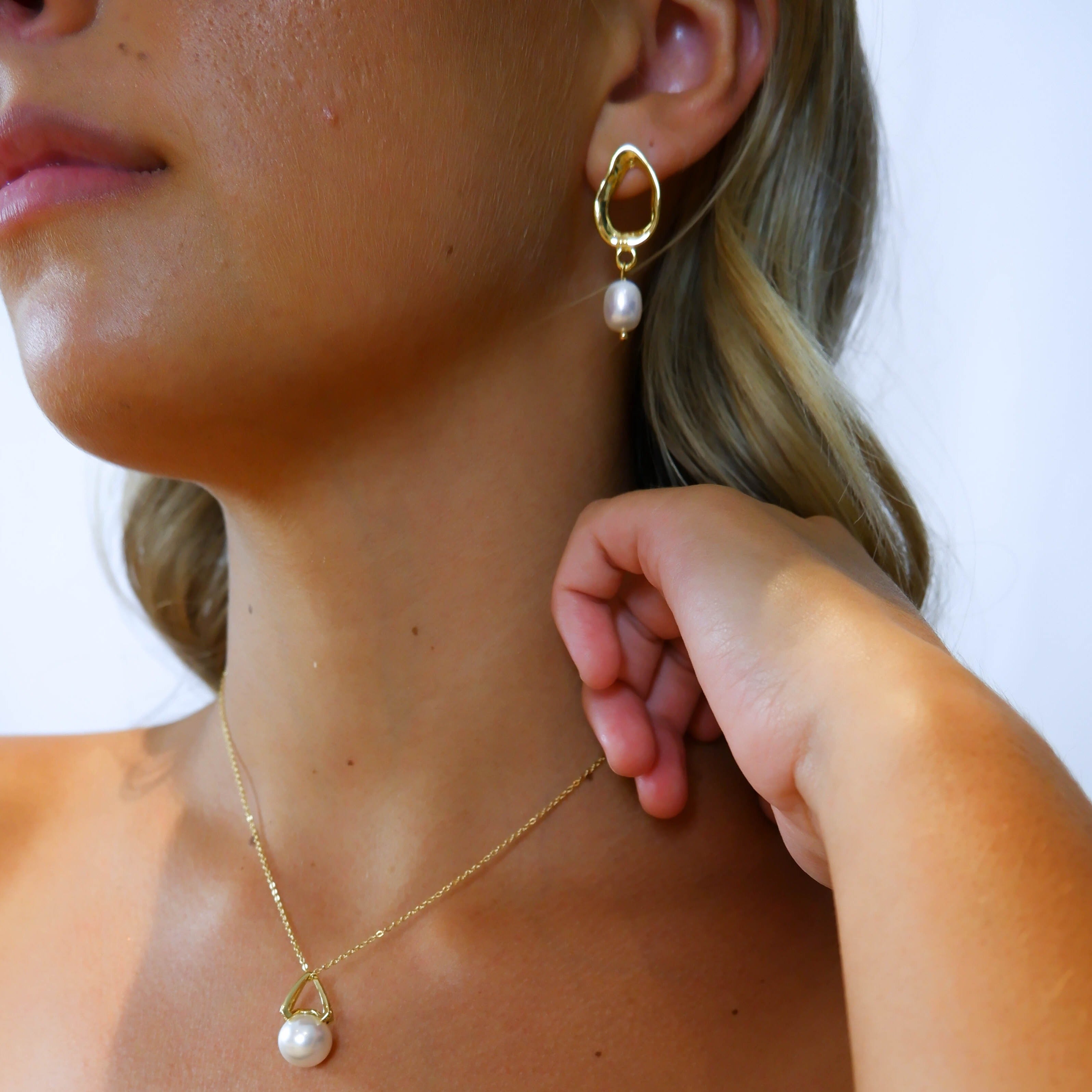 Close-up of model’s neck and ear wearing gold Sophia Earrings with a single pearl pendant necklace – hand brushing hair aside