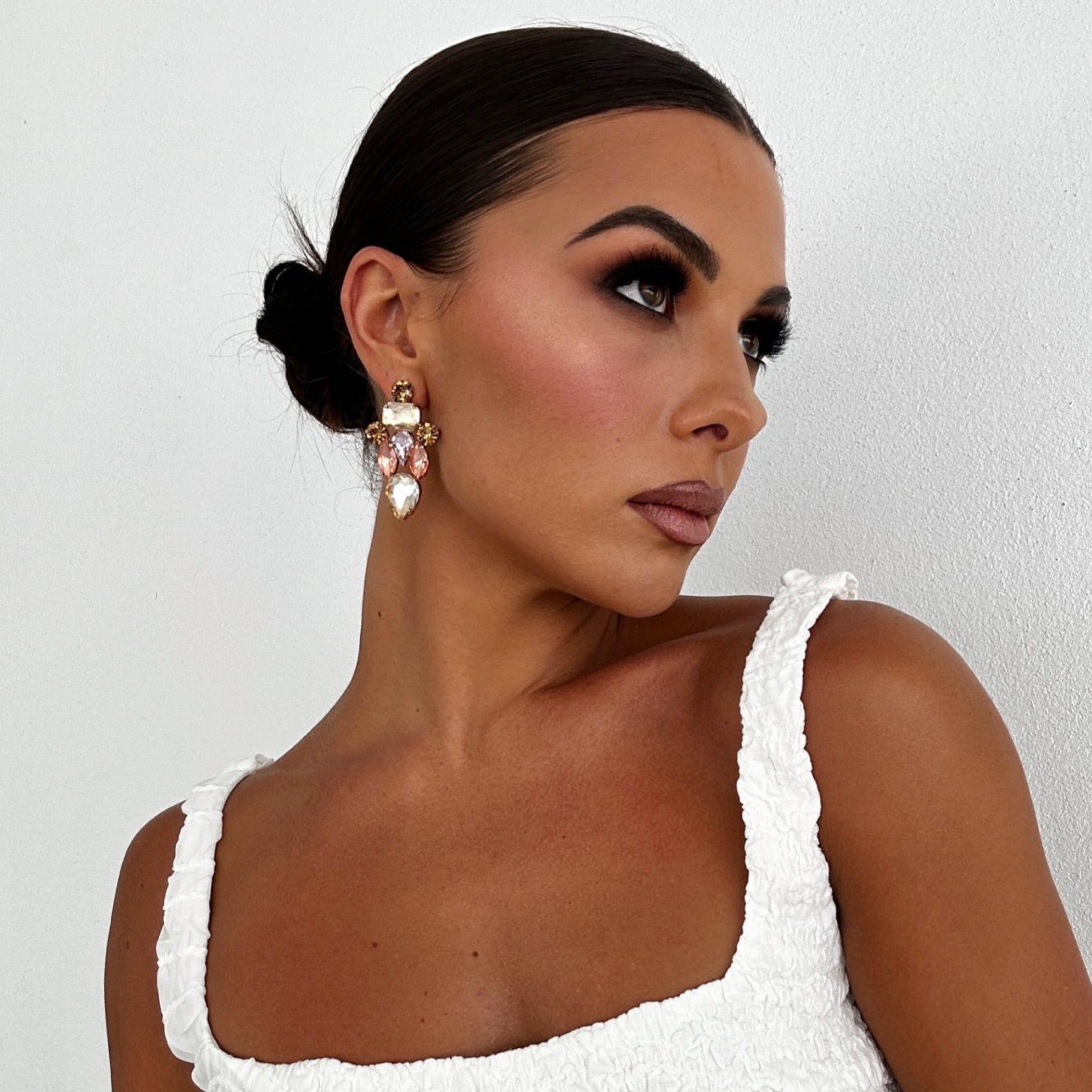 Woman with dark slicked-back hair wearing Ciara Earrings in champagne – styled with white textured top and bronzed makeup