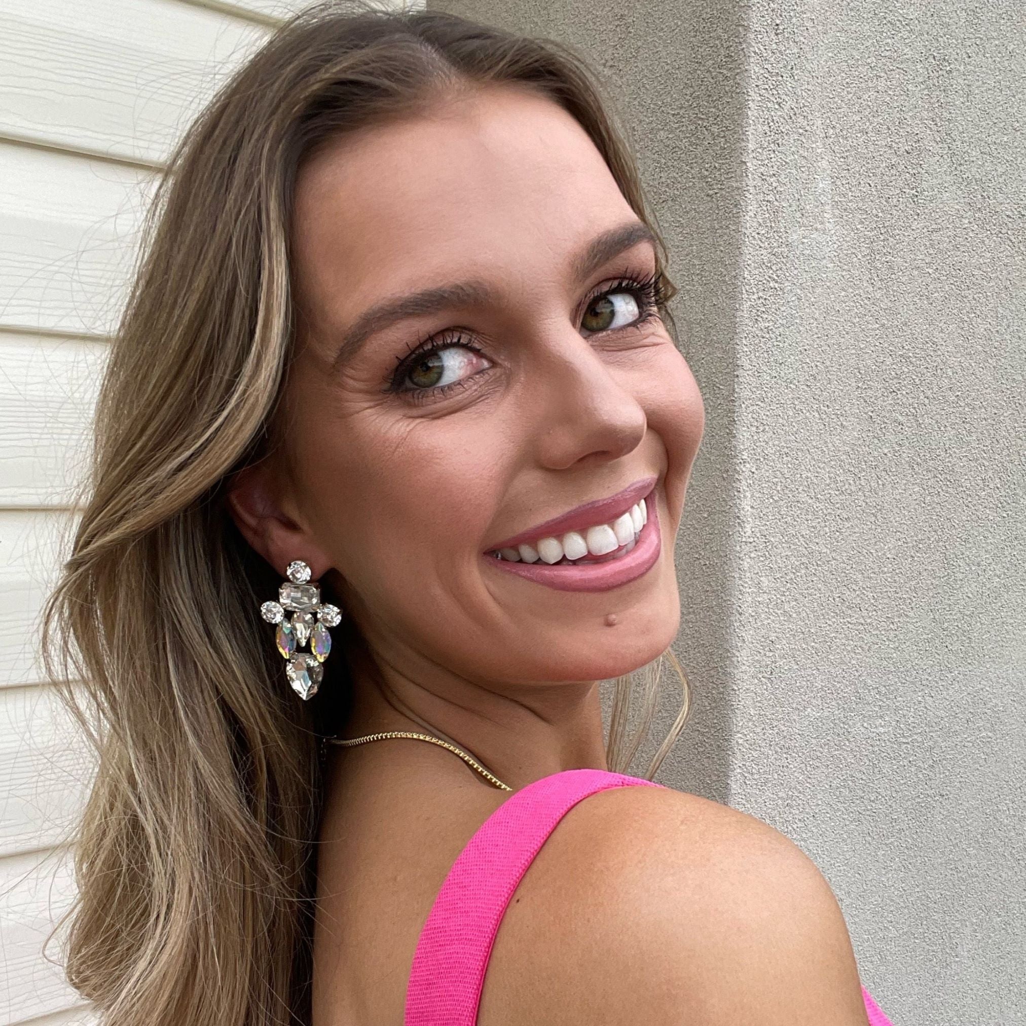 Woman smiling in pink dress wearing Ciara Earrings in silver and white crystal – soft glam look with long hair and natural light backdrop