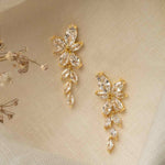 Gemma Earrings in gold – trailing cubic zirconia earrings resting on ivory cloth with delicate floral accents.