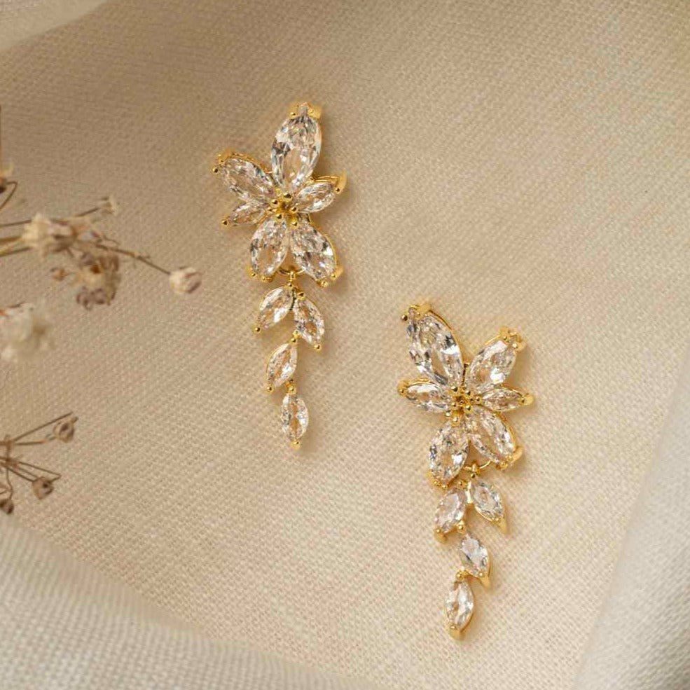 Gemma Earrings in gold – trailing cubic zirconia earrings resting on ivory cloth with delicate floral accents.