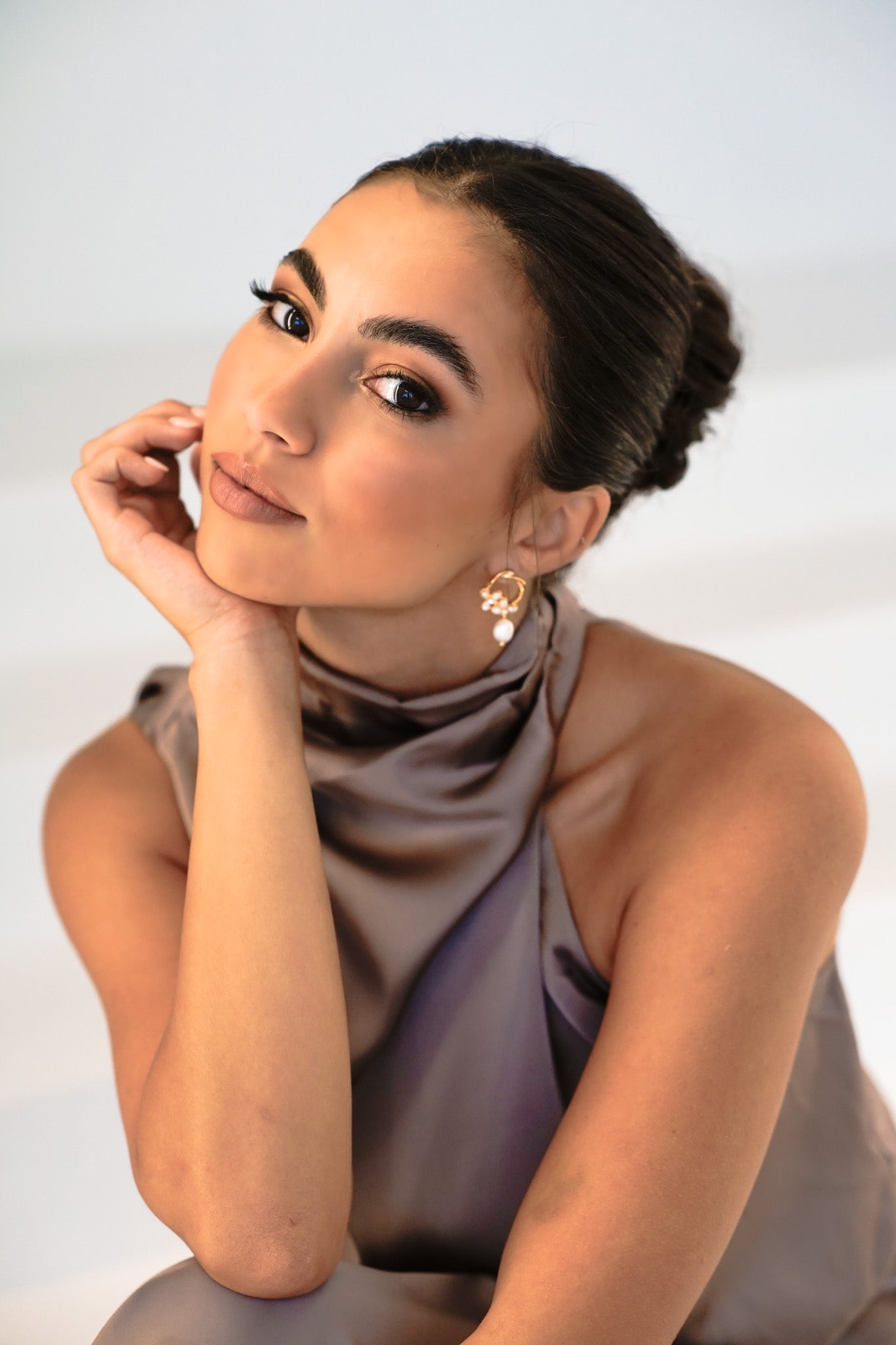 Bridesmaid wearing Jayda Earrings with dark hair in bun and purple halter dress – leaning head on palm, curious expression against white background